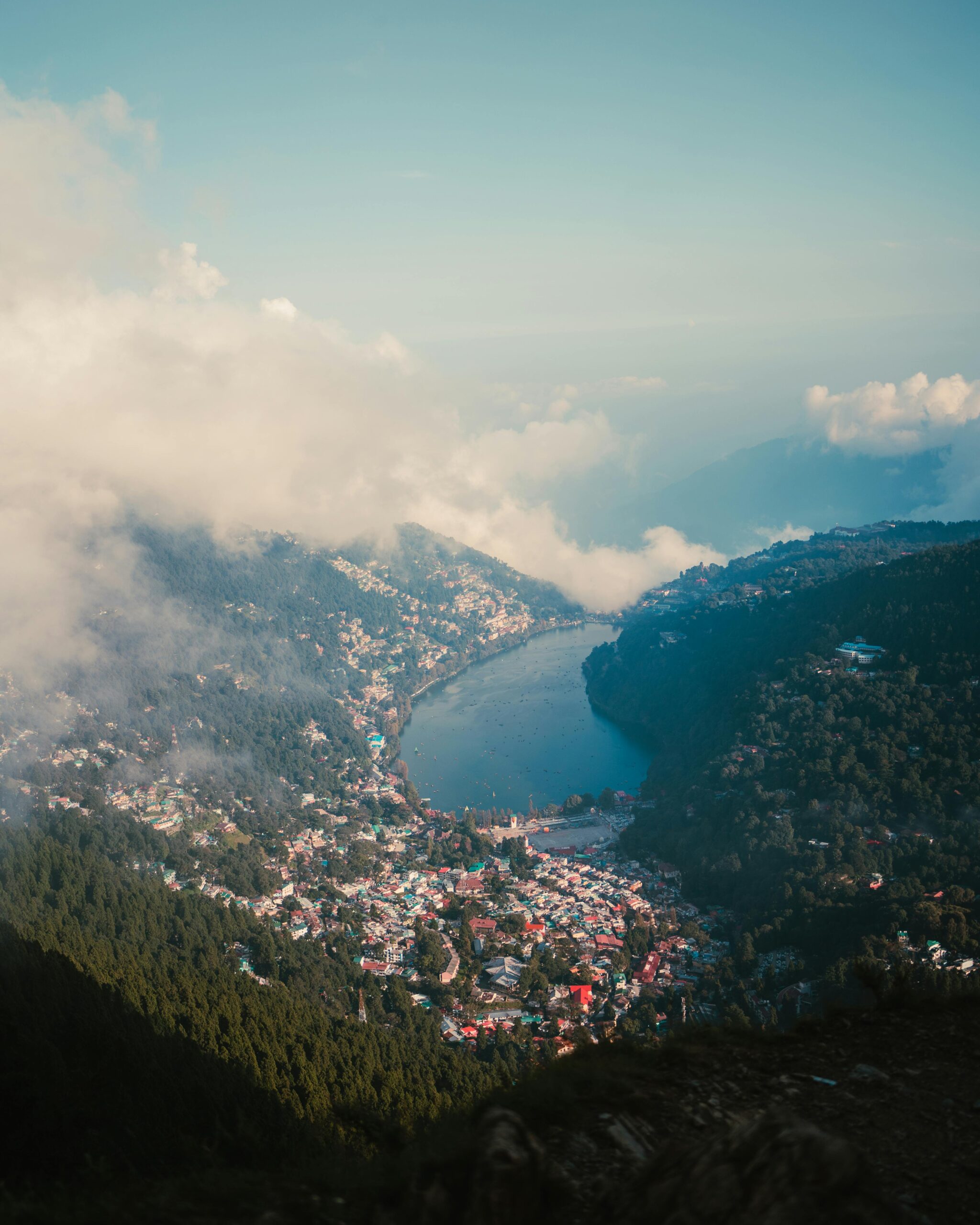 A breathtaking aerial view of Nainital Lake surrounded by mountains and clouds under a clear sky.